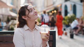 Happy young woman enjoying morning cold coffee drink with ice and smiling outdoors. Relaxing, taking a break. Girl sits on bench on city sunny street, drinking coffee to go. Town lifestyles outside video