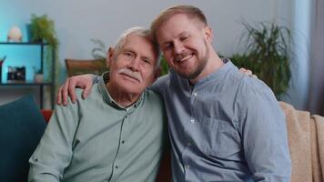 Portrait of senior grandfather with young adult man grandson smiling happy embracing hugging. Elderly father with son sitting on couch at home room. Male generations family. Bonding, good relationship video