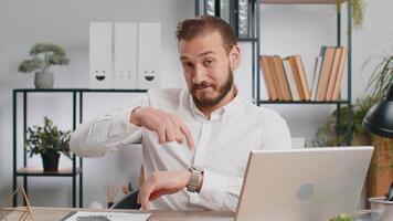 It is your time. Young middle eastern boss business man showing time on wrist watch clock, approve, pointing finger, time to start work, deadline. Freelancer guy at office workplace looking at camera video