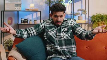 I dont know what to say. Confused indian handsome man feeling embarrassed about ambiguous question, having doubts, no idea, being clueless and uncertain at home. Hindu guy sits on sofa in living room video