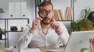 Investigator researcher scientist businessman working at home office holding magnifying glass near face looking into camera with big zoomed funny eye, searching, analysing. Professional freelancer man video