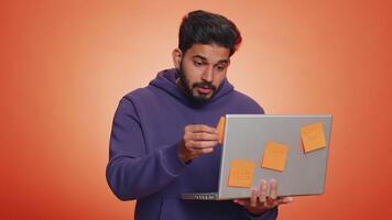 Exhausted indian freelancer man with pasted sticker notes use laptop computer having concentration problem creative crisis. Nervous hindu guy confused by big routine work loading on orange background video