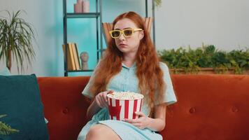 Excited redhead woman sitting on sofa eating popcorn and watching interesting tv serial, sport game, film, online social media movie content at home. Girl in 3D glasses enjoying domestic entertainment video