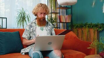 Man freelancer work with documents on home couch, reading information, business papers prepare financial report on laptop. Guy accountant analyzing charts and graph reports documents calculate taxes video