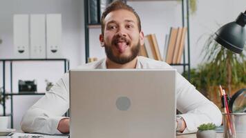 Lebanese businessman hiding behind laptop computer, making funny face, fooling around, putting his tongue out remote working disrespecting someone at office desk workplace. Professional freelancer man video