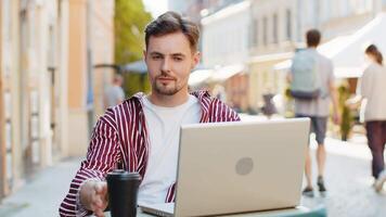 Man working study education online distant job with laptop in city sunny street cafe restaurant browsing website chatting outdoors during break, drinking coffee. Smiling guy tourist looking at camera video