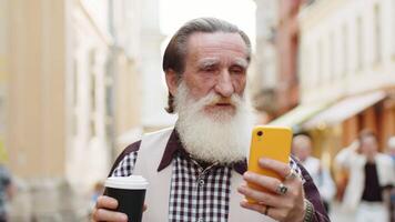 Happy senior old man use smartphone typing text messages browsing internet social media web app working chatting online enjoying morning to-go coffee hot drink. Grandfather tourist walking in street video