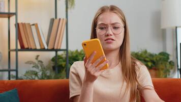 Portrait of young woman making phone call conversation with friend sitting on sofa. Happy excited girl enjoying mobile smartphone loudspeaker talking indoors at modern home living room apartment video