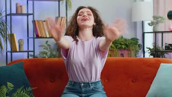 Hello. Young woman smiling friendly at camera and waving hands gesturing hello, hi, greeting or goodbye, welcoming with hospitable expression at home. Portrait of girl sitting on sofa in living room video