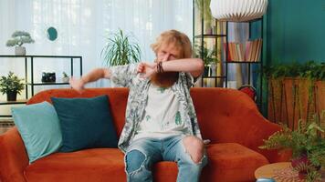 Trendy cheerful positive Caucasian redhead man having fun dancing, dabbing raising hands making dub winner dance gesture celebrating victory at home apartment. Happy bearded guy in living room on sofa video