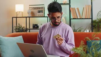Young indian man using credit bank card and laptop computer while transferring money, purchases online shopping, order food delivery. Portrait of hindu guy at home apartment living room on couch video