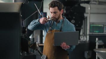 Caucasian man working in roasted coffee fabric inspecting roasting machine caffeine beans sniffing smell aroma checking typing laptop quality check holding computer engineer technologist male go away video