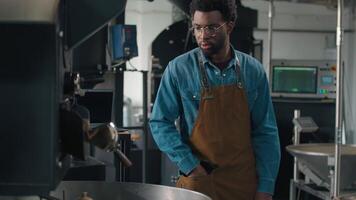 African American man working approach roasting machine pull lever coffee beans falling out roasted cocoa male roaster in apron guy at modern factory professional barista technician preparing caffeine video