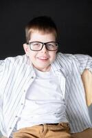 boy wearing reading glasses and hearing aid, white shirt sits on chair against black wall smiling looking at frame. photo