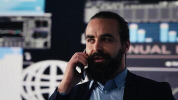 Businessman answering a smartphone call to evaluate digital analytics and corporate insight. Surrounded by finance data and trend reports, he strategizes leadership goals and projections. Camera A. video