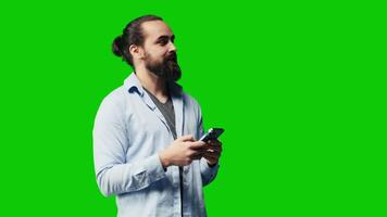 Casual stylish guy browsing his smartphone against green screen background, connected online on social network platforms or texting messages. Young adult focused on social media apps. Camera A. video