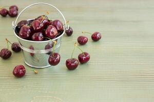 In frame on the left a small bucket with cherries, around scatterd cherries, on the right empty space on a light green background. Bucket of cherries. Horizontal. Daylight. photo
