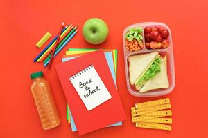 School supplies and lunch box with sandwiches, pieces of cucumbers and carrots, apricot, cherries, apple, bottle of juice on red background, back to school concept. Horizontal. Top view. photo