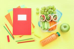 School supplies and lunch box with tasty rolls, cucumbers, carrots, grapes, apple, bottle of juice on light yellow background, back to school concept. Horizontal. Top view. photo