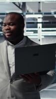 Vertical Technician walking in data center, configuring storage devices for minimal latency in AI workloads. African american employee using device in server farm to build neural networks, camera A video