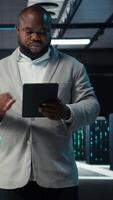 Vertical System administrator walking in data center analyzing infrastructure performance outputs on tablet. Server farm employee doing job tasks, finetuning supercomputers configurations, camera A video