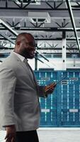Vertical Programmer in data center using tablet to manage software updates for system stability. African american man in server room optimizing performance with device, configuring infrastructure, camera A video