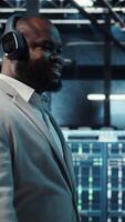 Vertical Happy worker wearing headphones walking in data center with hardware used to host websites. Cheerful IT expert listening music while overseeing dimly lit server farm rigs, camera A video