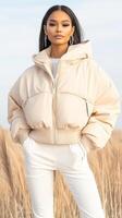 Woman stands wearing futuristic style. Neutral color puffed jacket outfit. Tall grass background, bright style. Outdoor fashion photo