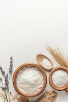 Wooden bowls contain white powder. Lavender flowers and dry wheat plants lie nearby on a plain surface with open space at top. photo