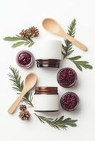 Jars with red jam sit near wooden spoons and evergreen branches with pine cones on a white surface viewed from above photo