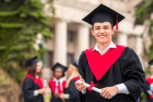 Handsome guy student in black and red graduation costume showing diploma and cheerfully smiling at camera, standing over chatting group of international students at university campus photo