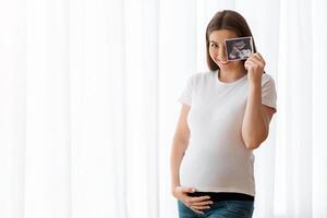 Happy Pregnant Woman With Ultrasound Scan Image Posing Near Window At Home, Cheerful Expecting Lady Embracing Her Belly And Smiling At Camera, Enjoying Healthy Pregnancy And Prenatal Time, Copy Space photo
