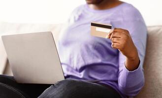 Making Financial Transaction. Closeup of unrecognizable black lady holding and showing credit card sitting on sofa with personal computer on her lap, blurred background. Online Shopping And Payment photo