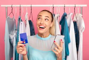 Retail And Purchase Concept. Excited stylish woman holding credit card and smartphone, looking up and thinking what to buy, ordering and buying clothes in online store via internet photo