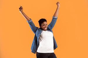 Joyful Oversized African American Woman Shaking Fists With Raised Hands Standing Posing Over Yellow Studio Background, Smiling To Camera. Joy, Positive Emotions Concept photo