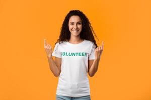 Smiling Volunteer Woman Pointing Fingers Upward Looking At Camera Standing In Studio Over Yellow Background. Look Up Concept, Volunteering Advertisement photo