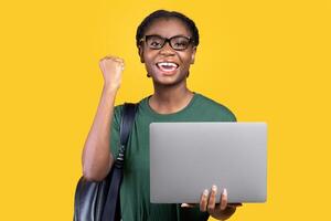 Joyful Black Female Student With Laptop Gesturing Yes After Successful Online Exam Standing Posing On Yellow Studio Background. Joy Of E-Learning, Educational Gadgets, Distant Study Concept photo