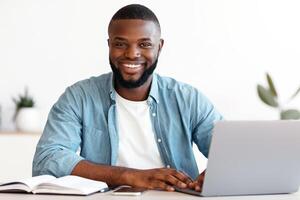 Portrait Of Smiling Successful African American Male Entrepreneur Posing At Workplace In Modern Office, Handsome Black Man Sitting At Desk With Laptop Computer And Looking At Camera, Copy Space photo
