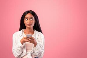 Young black lady holding smartphone and looking at blank space over pink studio background. Serious African American woman browsing web, chatting or checking email on mobile device photo