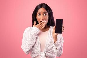 Pretty black lady showing smartphone with blank screen and covering her mouth in shock on pink studio background, mockup for design. Surprised young woman with empty cellphone promoting new mobile app photo