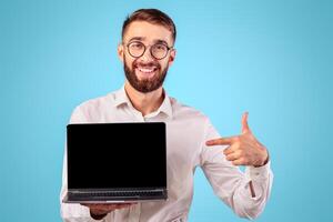 Positive young entrepreneur pointing at laptop computer with blank screen for website design on blue studio background. Happy millennial businessman with portable PC, space for mockup photo