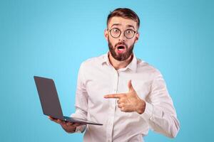 Shocked young man pointing at laptop computer with open mouth, surprised over web content on blue studio background. Millennial guy in formal wear receiving unbelievable offer photo