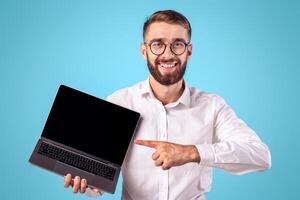 Happy millennial businessman pointing at laptop computer with mockup for website design on blue studio background. Smiling young male entrepreneur with portable PC, space for text photo