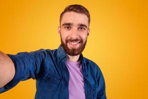Handsome young Caucasian guy taking selfie and smiling widely over orange studio background. Positive millennial man photographing himself on camera, making self portrait photo