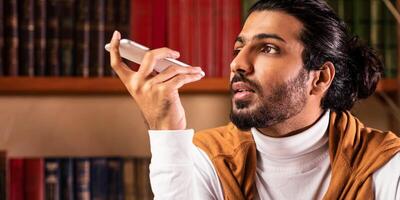 Indian Student Guy Using Voice Assistant On Smartphone Learning Sitting In University Library Indoors. Educational Application For Mobile Phone. Modern Education Concept. Selective Focus photo