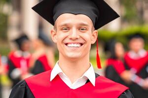 Handsome guy student posing in graduation costume, black and red robe and hat, cheerfully smiling at camera while having graduation ceremony at university campus, closeup portrait, copy space photo