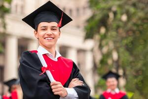 Happy young man student in graduation robe and hat showing diploma and cheerfully smiling at camera, standing over chatting group of multiracial students at university campus photo