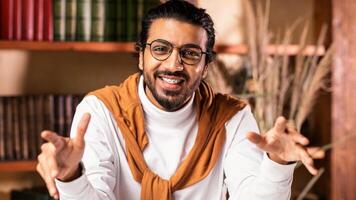 Indian Male Teacher Talking Smiling To Camera Explaining Lesson Teaching Online Sitting In Living Room Near Books Shelf At Home. Remote Tutoring And E-Teaching Concept. Selective Focus photo
