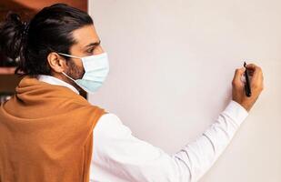 Teacher Man In Face Mask Teaching Writing On Blank White Blackboard Having Class Standing Indoors. Safe School And University Education, Tutoring, Distant And Online E-Teaching Concept. Mockup photo