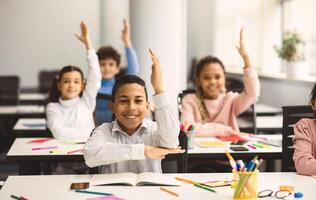 School Reopening, Back To Normal Lifestyle Concept. Portrait of excited little schoolboy raising hand up in the air to answer a question with all his classmates in blurred background, selective focus photo
